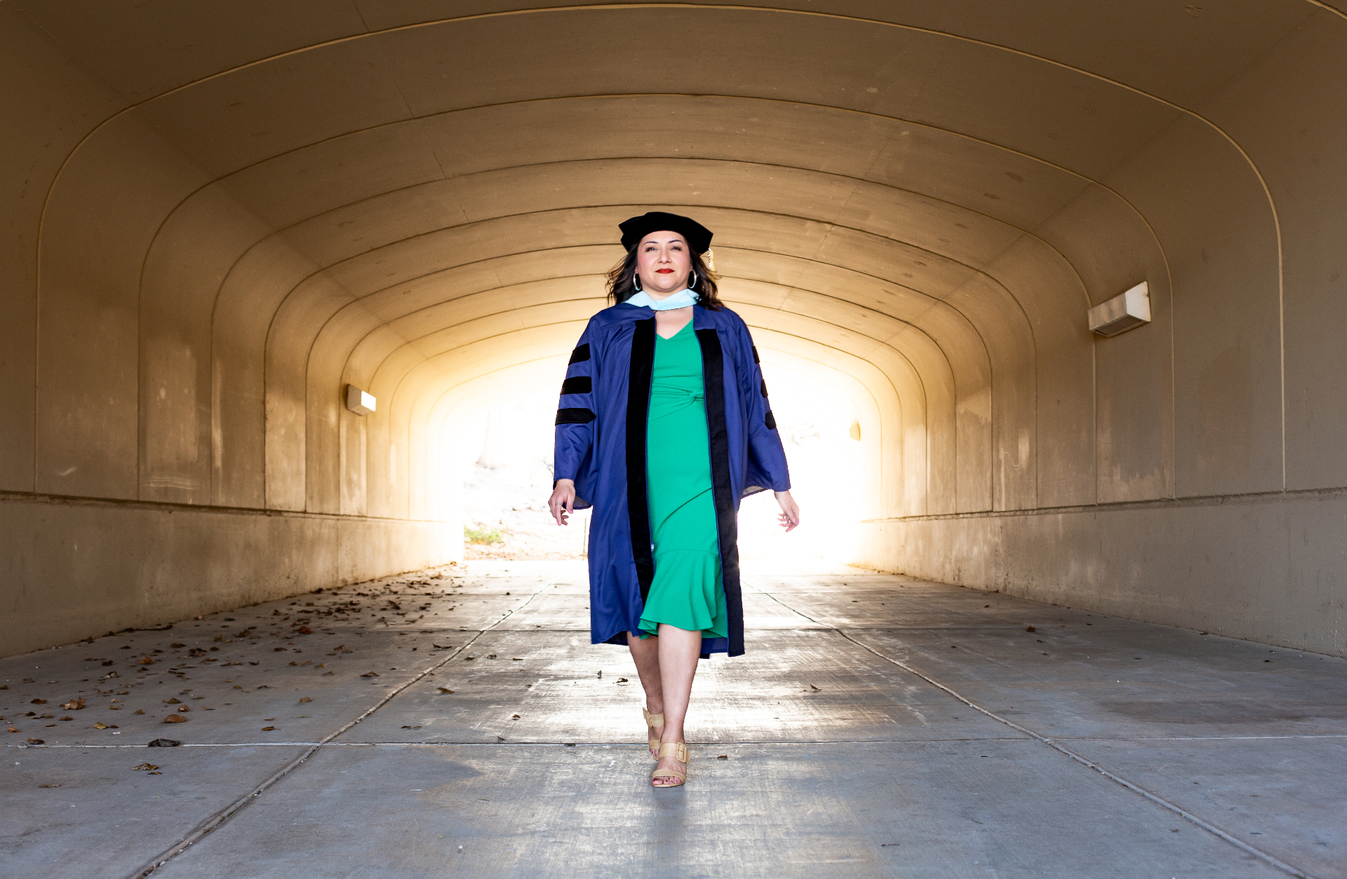 Dr. Martha A. Castillo walking forward in doctoral regalia through a light-filled tunnel